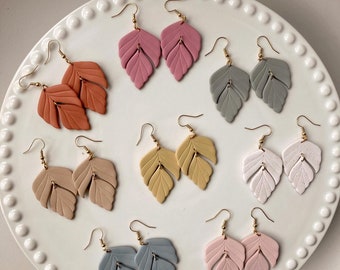May include: Assortment of eight pairs of handmade polymer clay earrings. Each pair features a double leaf design in a different color: coral, pink, gray, yellow, white, light blue, and light pink.