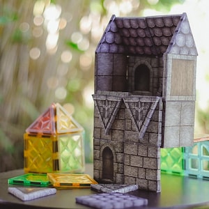 Puede incluir: Una exhibición de bloques de construcción coloridos, incluyendo una estructura gris con aspecto de piedra con ventanas arqueadas y un tejado de tejas. Otros bloques son transparentes y vienen en varios colores, como amarillo, verde y naranja. Los bloques están dispuestos sobre una superficie oscura.