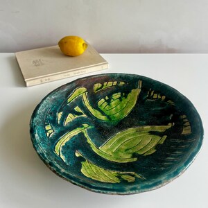 May include: A round ceramic bowl with a dark teal and deep blue glaze featuring hand-painted light green leaf motifs. The bowl has a textured, uneven rim and a glossy finish. A yellow lemon rests on a beige book in the background.
