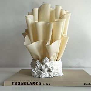 May include: A table lamp featuring a white sculpted base with a cherub figure playing a flute, surrounded by draped fabric. The lamp shade consists of tall, cream-colored, vertically pleated fabric panels that fan outward in a ruffled design.
