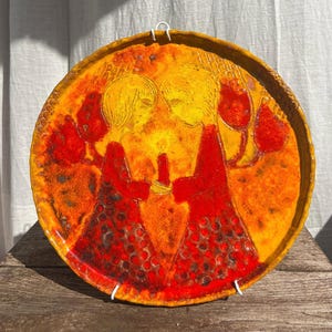 Può includere: Un piatto da parete rotondo in ceramica con una vivace smaltatura arancione e gialla. Il design raffigura due figure l'una di fronte all'altra che tengono una candela accesa. Le figure indossano abiti rossi a motivi e lo sfondo presenta forme floreali rosse stilizzate.
