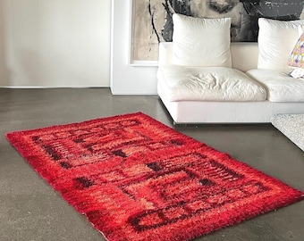 May include: A rectangular shag rug featuring a vibrant red base color with darker red and black geometric patterns. The rug has a thick, textured pile surface and is positioned flat on a smooth, grey floor surface in a room setting.