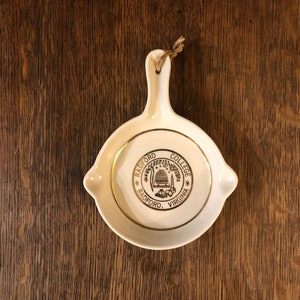 May include: White ceramic trinket dish with a gold rim and a gold logo that reads "Radford College Radford, Virginia". The logo features a building with a beehive on top.