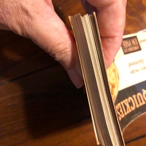 K&ouml;nnte beinhalten: Nahaufnahme des Buchr&uuml;ckens eines Taschenbuchs mit cremefarbenem Einband. Das Buch wird in einer Hand gehalten und die Seiten sind sichtbar.