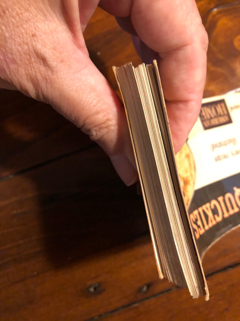 K&ouml;nnte beinhalten: Nahaufnahme des Buchr&uuml;ckens eines Taschenbuchs mit cremefarbenem Einband. Das Buch wird in einer Hand gehalten und die Seiten sind sichtbar.