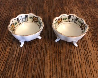 May include: Two small white ceramic salt cellars with three legs each. The cellars have a scalloped rim and are decorated with a floral pattern in shades of yellow, green, and brown.