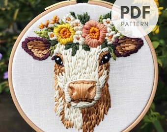 Könnte beinhalten: Gesticktes Kuhgesicht mit Blumenkranz in einem Holzreifen. Die Kuh hat braunes und weißes Fell, mit einem detaillierten Blumenkranz aus gelben, rosa und weißen Blumen. Der Text "PDF PATTERN" befindet sich in einem weißen Kreis.
