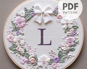 Pode incluir: Arte de bordado em aro com uma coroa floral com flores rosa, brancas e roxas, folhas verdes e um laço de cetim branco. A letra "L" é bordada no centro com fio roxo. O aro é de madeira e tem um fundo de tecido branco. O texto "PDF PATTERN" é exibido no canto superior direito da imagem.