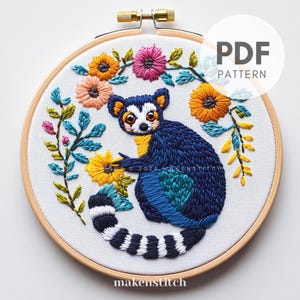 May include: Embroidered hoop art featuring a blue lemur with a white and black striped tail, surrounded by colourful flowers. The hoop is made of wood and has a white background.