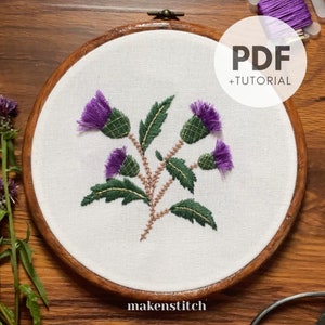 May include: Embroidered thistle flower design in a wooden embroidery hoop. The design features purple thistles with green leaves and brown stems. The hoop has the text "makenstitch" at the bottom.