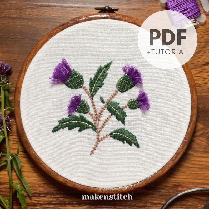 May include: Embroidered thistle flower design in a wooden embroidery hoop. The design features purple thistles with green leaves and brown stems. The hoop has the text "makenstitch" at the bottom.