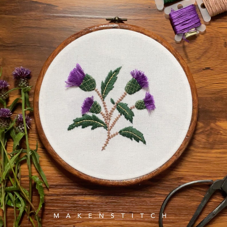 May include: Embroidered purple thistle flowers with green leaves on a white fabric in a wooden embroidery hoop.