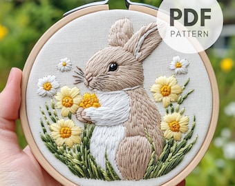 Puede incluir: Arte de bordado en aro con un conejo marrón con pelaje blanco que sostiene una flor amarilla, rodeado de margaritas blancas y amarillas y hierba verde. El aro es de madera y tiene un acabado natural. El texto "PDF PATTERN" es visible en la esquina superior derecha de la imagen.