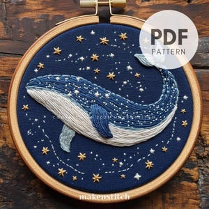 May include: Embroidered hoop art featuring a blue whale swimming through a starry night sky. The whale is outlined in white thread and has a blue and white gradient. The stars are gold and white.
