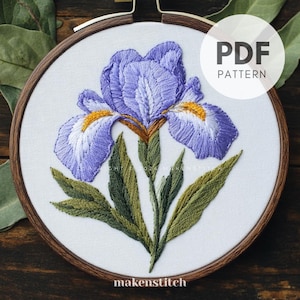 May include: A PDF pattern for embroidery of a purple iris flower with green leaves. The pattern is displayed on a white background in a wooden embroidery hoop. The text "PDF PATTERN" is displayed in the upper right corner of the image.