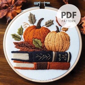 Peut inclure: Art de broderie en cercle représentant deux citrouilles et une pile de livres. Les citrouilles sont orange et brun avec des feuilles vertes. Les livres sont bruns, orange et crème avec des détails noirs et dorés. Le texte "PDF PATTERN" est dans le coin supérieur droit de l'image.