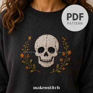 Könnte beinhalten: Ein dunkel anthrazitfarbenes Sweatshirt mit einer zentralen Stickerei eines cremefarbenen menschlichen Schädels, flankiert von zwei symmetrischen Zweigen mit kleinen orangefarbenen Blüten und grünen Blättern. Ein weißes Abzeichen zeigt PDF PATTERN.