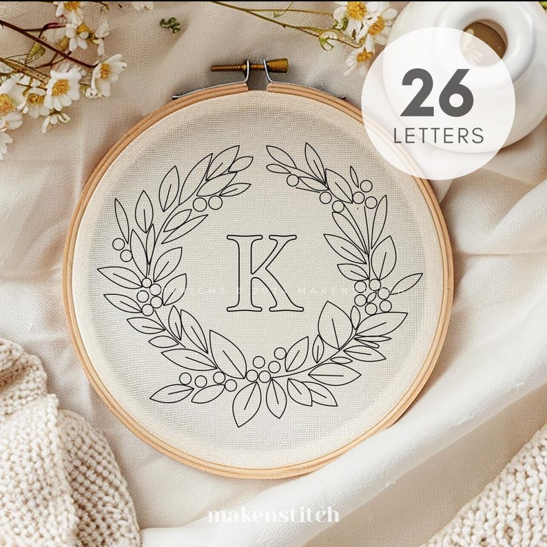 Pode incluir: Um aro de bordado branco com um contorno preto de uma coroa de folhas e a letra K no centro. O aro est&aacute; sobre um fundo de tecido branco. O texto "26 Letters" &eacute; vis&iacute;vel no canto superior direito da imagem.