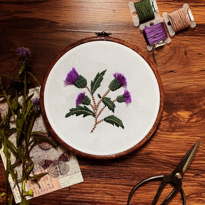May include: A close-up of a circular embroidery hoop with a white fabric background. The fabric features a purple and green embroidered design of a thistle flower. The hoop is on a wooden surface.