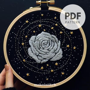 Könnte beinhalten: Gesticktes Stickrahmen-Kunstwerk mit einer grauen Rose, goldenen Sternen und gepunkteten Linien auf schwarzem Hintergrund. Der Text "PDF PATTERN" befindet sich in einem weißen Kreis. Der Holzrahmen wird von einer Hand gehalten.