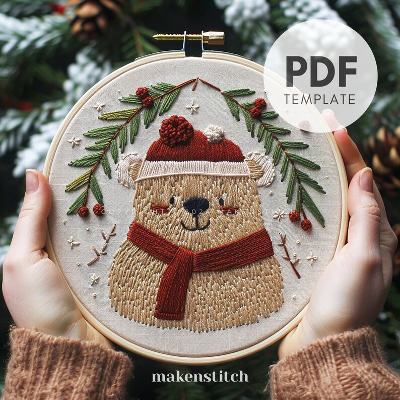 May include: Embroidered hoop art featuring a brown bear wearing a red Santa hat and scarf, surrounded by green pine branches with red berries and white snowflakes. The hoop is made of wood and has a white background. The text "makenstitch" is printed below the hoop.