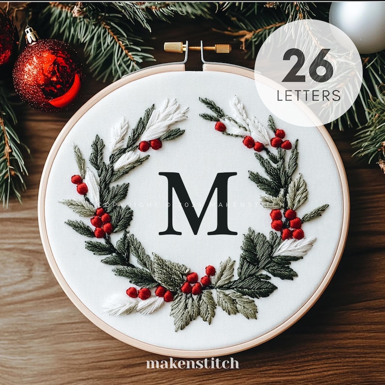 Pode incluir: Um aro de bordado branco com um design de coroa de Natal com folhas verdes e brancas, bagas vermelhas e a letra "M" em preto. O aro &eacute; de 26 letras e tem uma moldura de madeira.