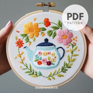 Può includere: Ricamo floreale in un telaio di legno. Il disegno presenta una teiera azzurra con fiori colorati, circondata da fiori arancioni, rosa e gialli e foglie verdi. Il testo "PDF PATTERN" è visibile.