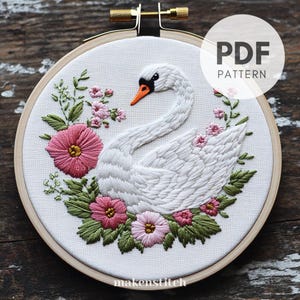Pode incluir: Design de cisne bordado em um bastidor de madeira. O cisne branco tem um bico laranja e é cercado por flores rosa e folhas verdes. O texto "PDF PATTERN" está em um círculo branco. O texto "makenstitch" está na parte inferior.