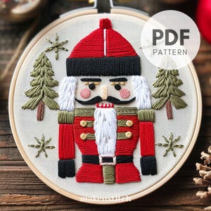 Peut inclure: Un modèle PDF pour un projet de broderie de casse-noisette de Noël. Le motif représente un casse-noisette rouge et vert avec une barbe blanche et un chapeau noir, debout entre deux arbres verts. Le motif comprend le texte "PDF PATTERN".
