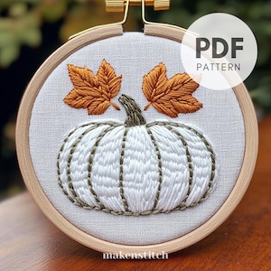 Peut inclure: Citrouille blanche avec une tige verte et deux feuilles orange brodées sur un tissu de lin blanc dans un cercle de broderie en bois. Le texte "PDF PATTERN" est dans un cercle blanc avec un contour noir.