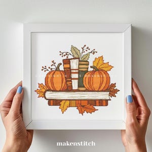 May include: A framed cross-stitch design showing a stack of books, two orange pumpkins, and autumn leaves. The design is in a white frame and held by hands with blue nail polish. The text "makenstitch" is at the bottom.