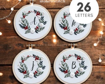 Pode incluir: Quatro aros de bordado com tecido branco e um design de coroa de Natal. Cada aro apresenta uma letra diferente do alfabeto, A, B, J e K. Os aros estão sobre uma superfície de madeira com luzes de corda. O texto "26 LETRAS" está em um círculo acima dos aros.