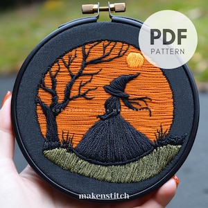 May include: Embroidered hoop art featuring a witch silhouette in a black dress and hat, standing in front of a large orange moon with a tree behind her. The background is a dark green field.