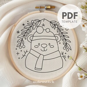 May include: Black and white line art embroidery pattern of a bear wearing a hat and scarf with a wreath of holly berries and leaves around the head. The pattern is on a white background and says "makenstitch" at the bottom.