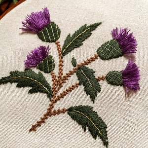 May include: Embroidered design of three purple thistle flowers with green leaves and brown stems on a white fabric background.