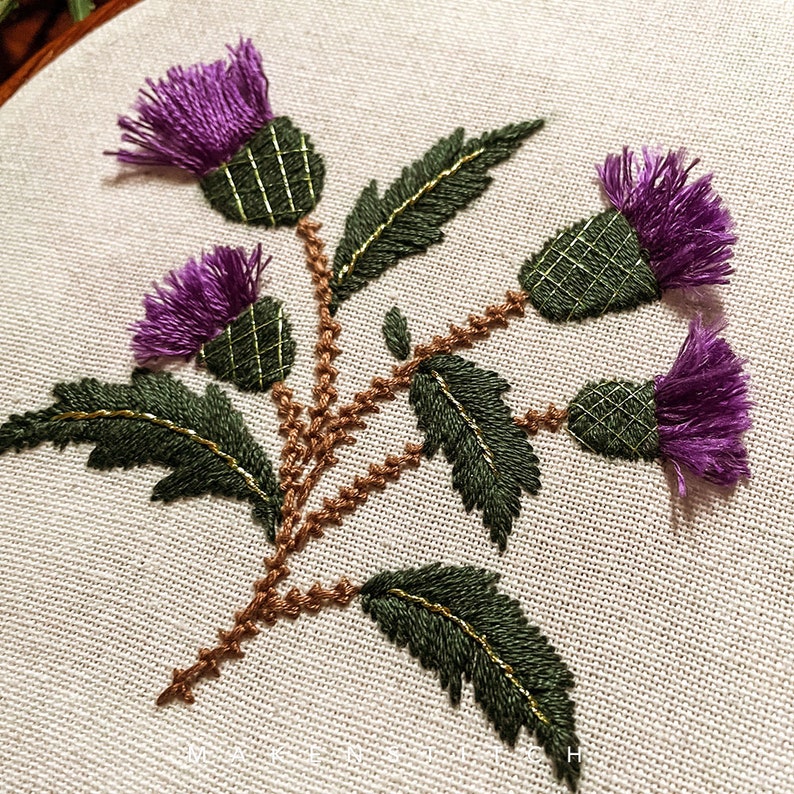 May include: Embroidered design of three purple thistle flowers with green leaves and brown stems on a white fabric background.