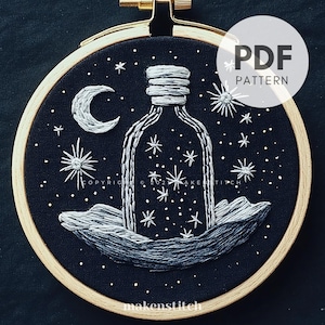 May include: A black embroidery hoop with a white embroidered design of a bottle filled with stars, a crescent moon, and waves. The text "PDF PATTERN" is in the top right corner of the hoop.