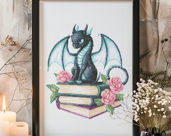 May include: Framed cross-stitch art featuring a black dragon with teal wings and accents, perched atop a stack of books adorned with pink roses. The artwork is set against a neutral background, with the text "Copyright 2025 makenstitch" visible.
