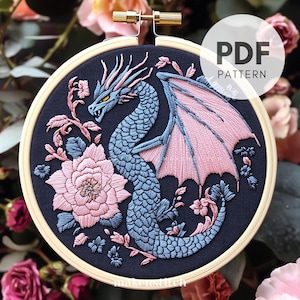 Könnte beinhalten: Gesticktes Drachendesign in einem Holzreifen. Der Drache ist blau mit rosa Flügeln und Akzenten, umgeben von rosa und blauen Blumenstickereien auf marineblauem Hintergrund. Der Text "PDF PATTERN" ist sichtbar.