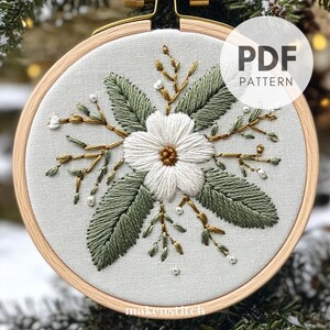 May include: A white flower with green leaves and gold stems is embroidered on a white fabric in a wooden embroidery hoop. The text "PDF PATTERN" is on the hoop.