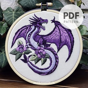 Könnte beinhalten: Gesticktes Drachendesign in Lilatönen, mit weißem Hintergrund, in einem hölzernen Stickrahmen dargestellt. Der Drache ist von lila Blumen und grünen Blättern umgeben. Der Text "PDF PATTERN" befindet sich in einem weißen Kreis.