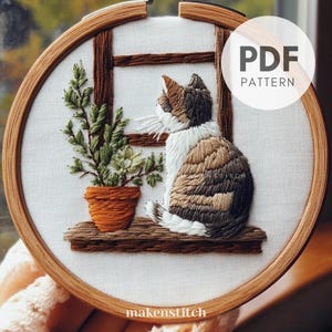 May include: Embroidered hoop art featuring a calico cat sitting on a windowsill with a potted plant. The cat is looking out the window. The hoop is made of wood and has a white fabric background. The text "PDF PATTERN" is in the upper right corner of the image.