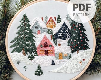 Peut inclure: Art de broderie sur un cercle représentant une scène de village d'hiver avec des maisons recouvertes de neige, des sapins et un paysage enneigé. Le cercle est en bois et a un fond blanc. Le texte "PDF PATTERN" est visible dans le coin supérieur droit de l'image.