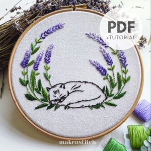 Könnte beinhalten: Ein Stickmuster eines schlafenden weißen Katers inmitten von Lavendelblüten, in einem Stickrahmen dargestellt. Das Muster beinhaltet ein PDF-Tutorial. Der Text "makestitch" befindet sich am unteren Rand des Rahmens.