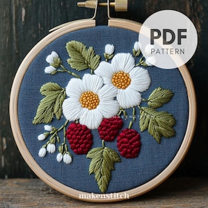 Puede incluir: Diseño floral bordado en un aro de madera. Presenta flores blancas con centros amarillos, bayas rojas y hojas verdes sobre un fondo azul. El texto "PDF PATTERN" está en un círculo blanco. El texto "makenstitch" está en la parte inferior.