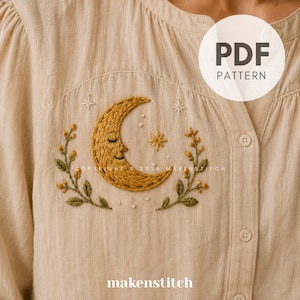 May include: A beige button-down shirt featuring an embroidered design of a golden crescent moon with a face, surrounded by green leafy branches and small white stars. The embroidery is centered on the chest area of the garment.