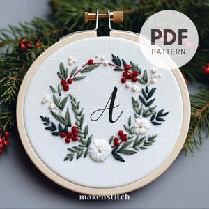 Pode incluir: Um aro de bordado branco com um design de coroa floral verde e vermelha. A coroa é feita de folhas, bagas e flores. A letra "A" é bordada no centro da coroa. O aro é etiquetado com "makestitch" na parte inferior.