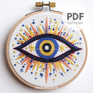 Peut inclure: Un modèle PDF pour une broderie d'œil colorée. L'œil est bleu, jaune et noir avec un rayonnement de soleil orange et jaune autour. Le motif est sur un tissu blanc dans un cerceau de broderie en bois.
