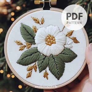 May include: Embroidered floral design in a wooden hoop. The white flower has a golden center, surrounded by green leaves and golden accents. The text "PDF PATTERN" is in a white circle. The text "makenstitch" is at the bottom.