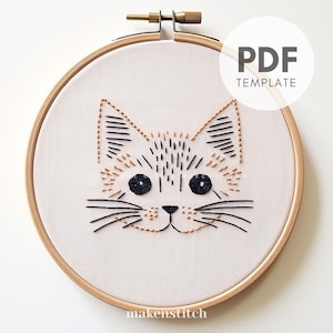 May include: Embroidered cat face design in a wooden embroidery hoop. The cat has black and orange thread details and black eyes.